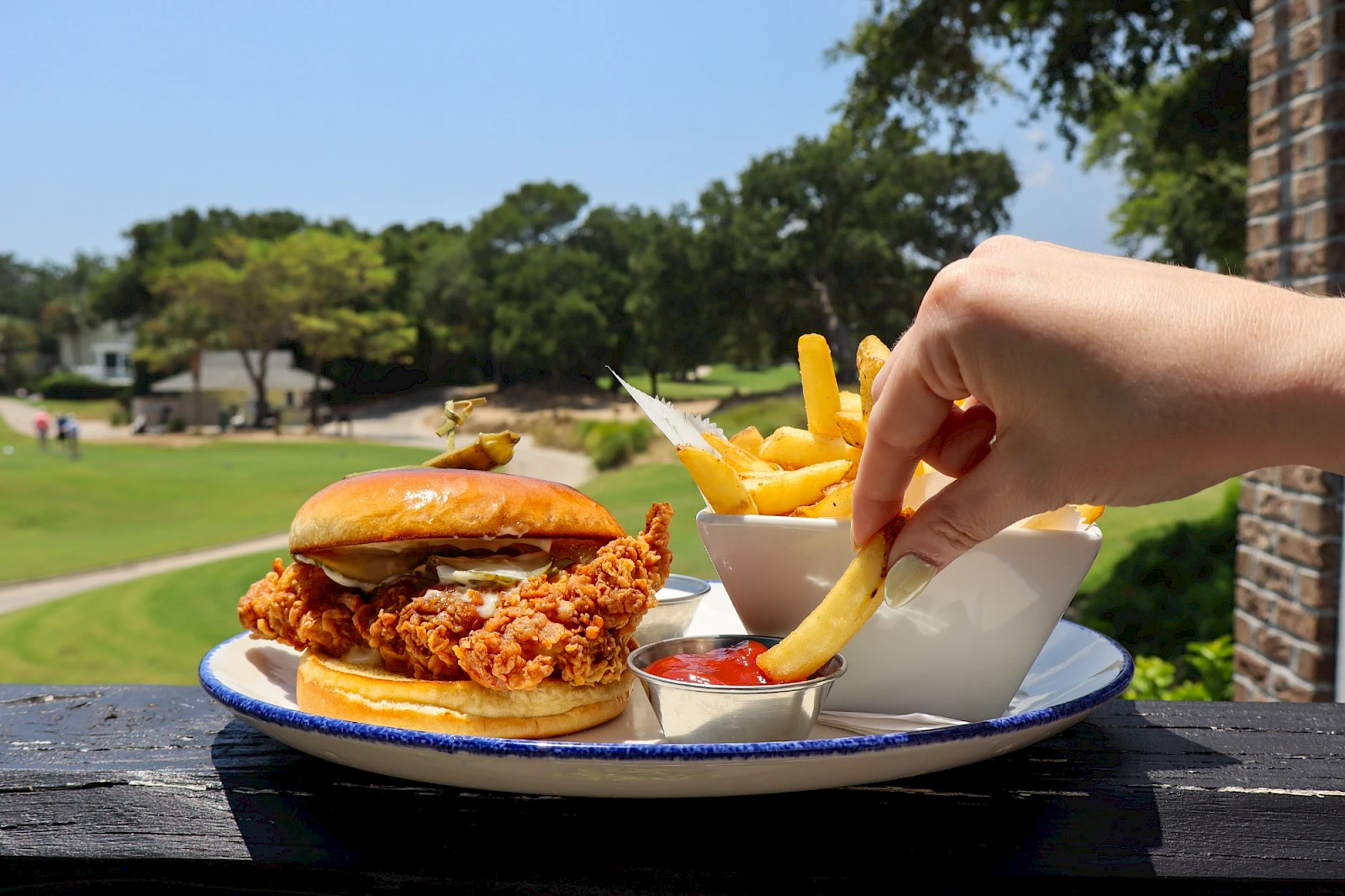 A crispy fried chicken sandwich with fries and dipping sauces on a plate, outdoors on a sunny day.