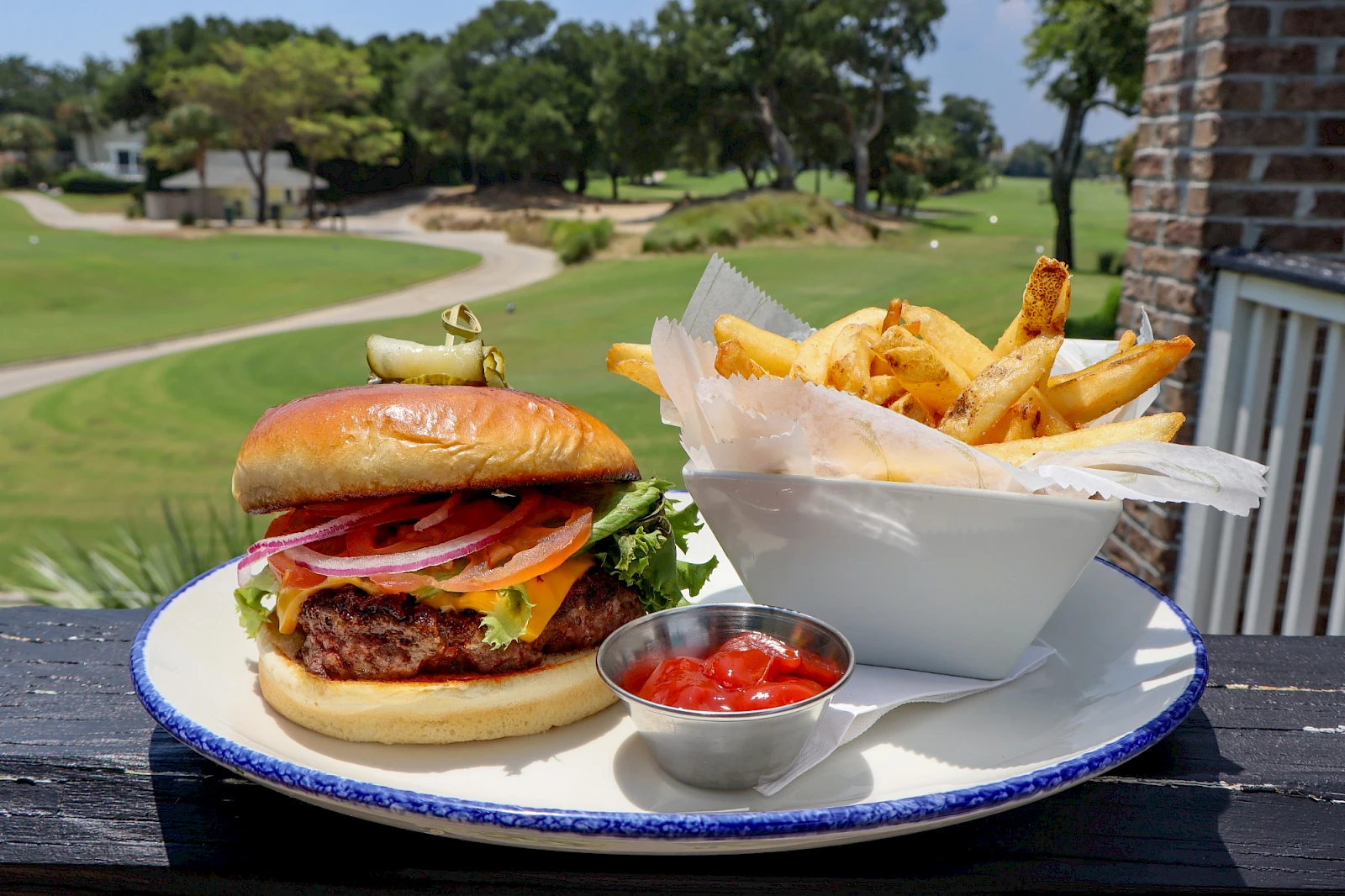 A juicy burger with bacon and tomato, crisp fries in a white pouch, and a small cup of ketchup on a blue-rimmed plate, outdoors.