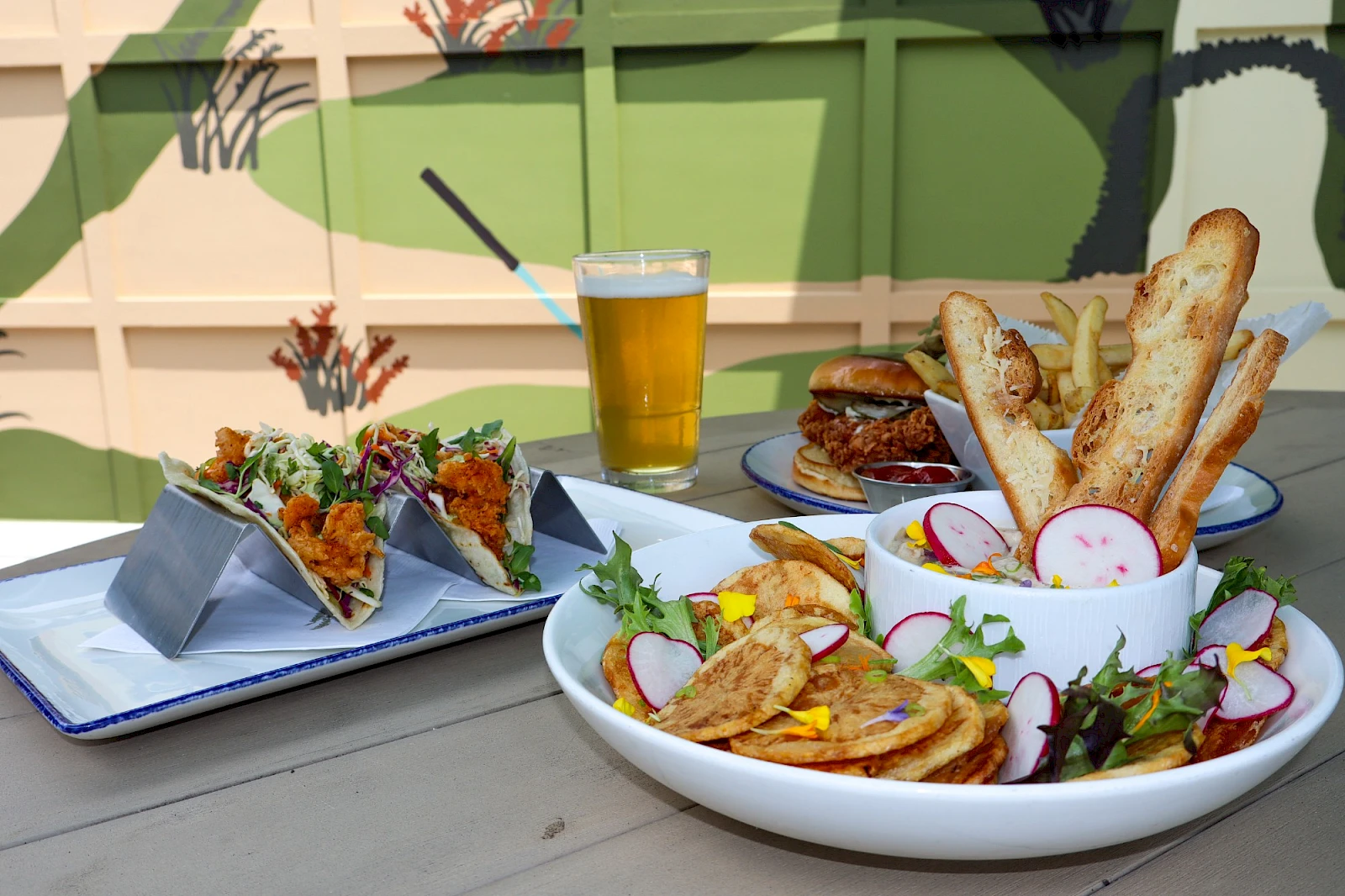 A colorful outdoor meal: a white bowl of nachos with veggies, a side of baguette slices, radish, greens; a small plate of tacos or sliders in back; a glass of beer in the center.