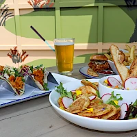 A colorful outdoor meal: a white bowl of nachos with veggies, a side of baguette slices, radish, greens; a small plate of tacos or sliders in back; a glass of beer in the center.