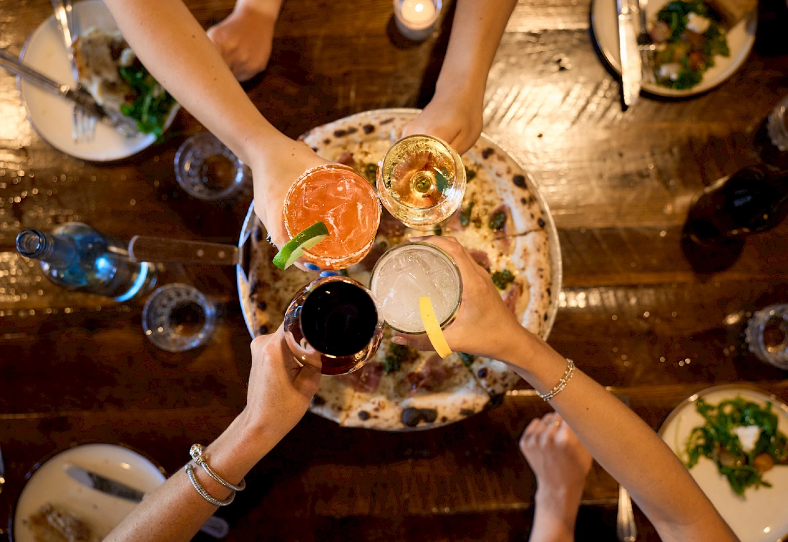 A group of friends cheers with colorful cocktails around a shared plate on a rustic wooden table, hands raised in celebration.