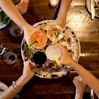 A group of friends cheers with colorful cocktails around a shared plate on a rustic wooden table, hands raised in celebration.