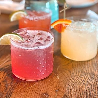 Three colorful cocktails on a wooden table, with garnishes of lemon and orange slices, a small plant in the background, casual bar vibe.