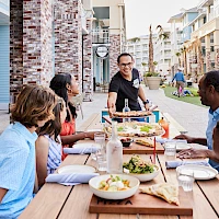 A group of diverse people sits at a long outdoor table on a sunny street, sharing a meal with plates, bread, and drinks while a server stands nearby.