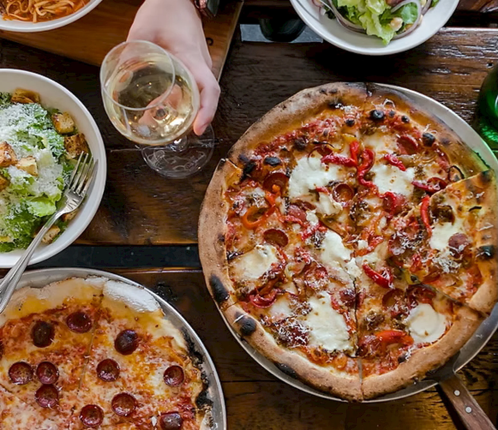 A table with two pizzas (pepperoni and a veggie/white cheese), a glass of white wine, a salad, and a hand holding the glass, all on a wooden table.