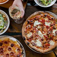 A table with two pizzas (pepperoni and a veggie/white cheese), a glass of white wine, a salad, and a hand holding the glass, all on a wooden table.