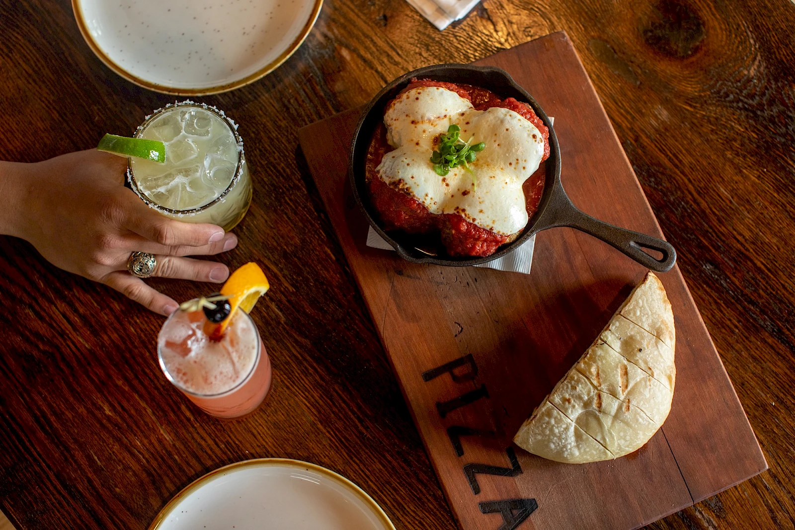 A sizzling skillet with melted cheese-topped dish, a side of flatbread on a wooden board labeled “PIZZA,” and two colorful cocktails beside a hand on a wooden table.