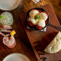 A sizzling skillet with melted cheese-topped dish, a side of flatbread on a wooden board labeled “PIZZA,” and two colorful cocktails beside a hand on a wooden table.
