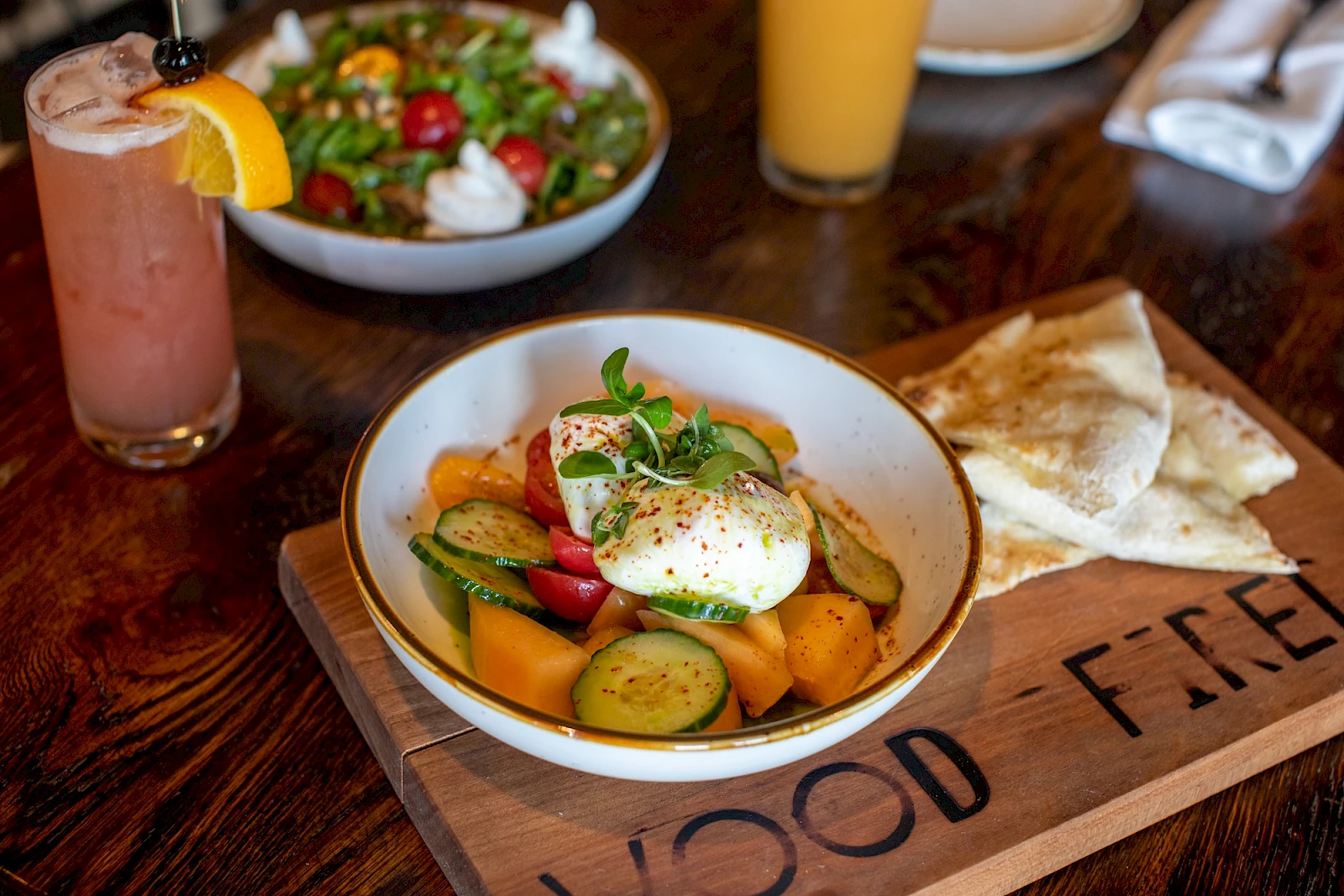 Colorful vegetable ratatouille in a bowl with a poached egg on top, grilled bread on the side, fresh salad in the background, and a pink smoothie to the left.