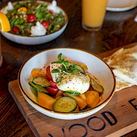 Colorful vegetable ratatouille in a bowl with a poached egg on top, grilled bread on the side, fresh salad in the background, and a pink smoothie to the left.