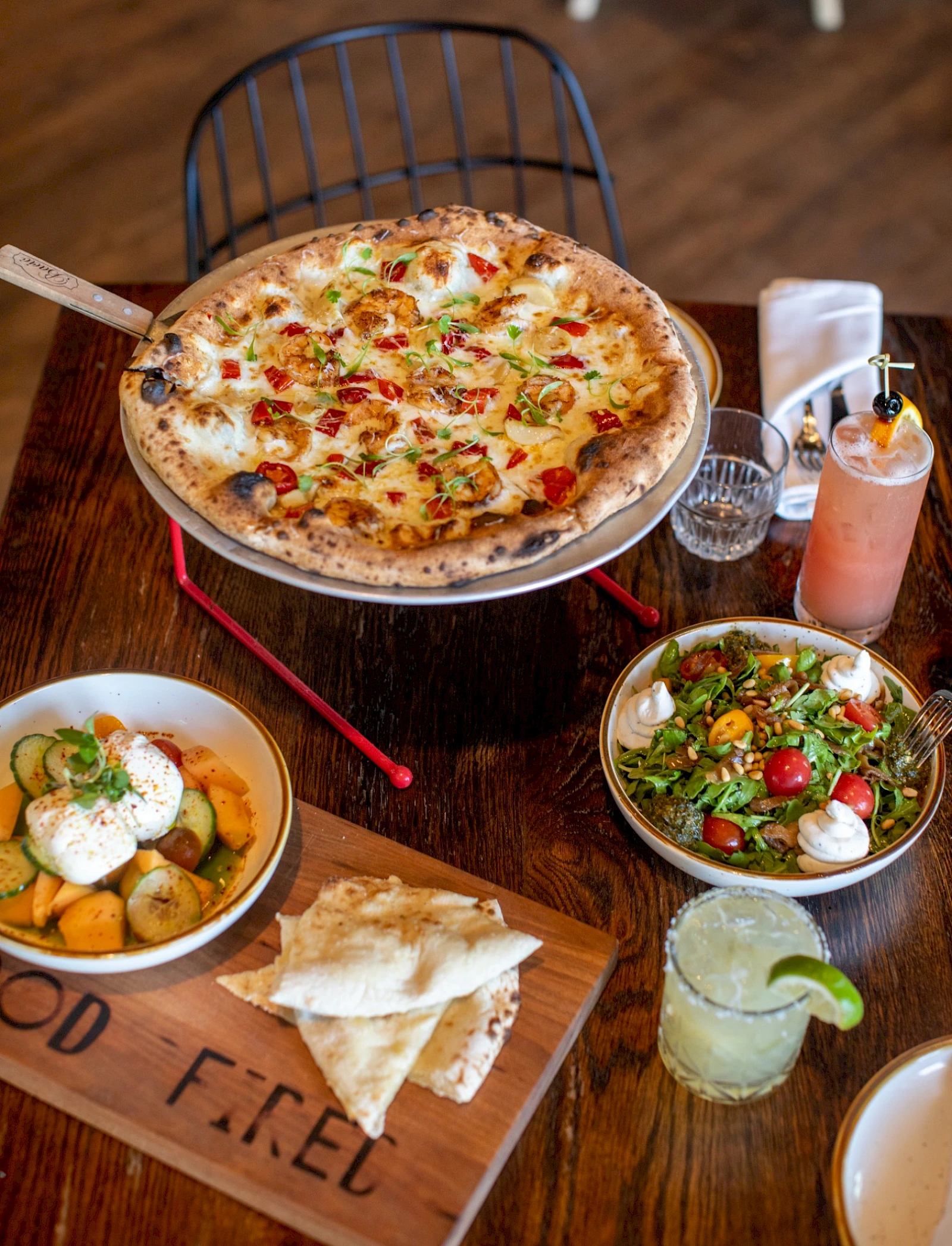 A hot cheese pizza with toppings on a round tray, plus garlic bread, potato sides, fresh salad, and a lime-garnished drink on a wooden table.