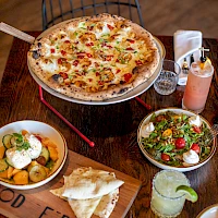 A hot cheese pizza with toppings on a round tray, plus garlic bread, potato sides, fresh salad, and a lime-garnished drink on a wooden table.