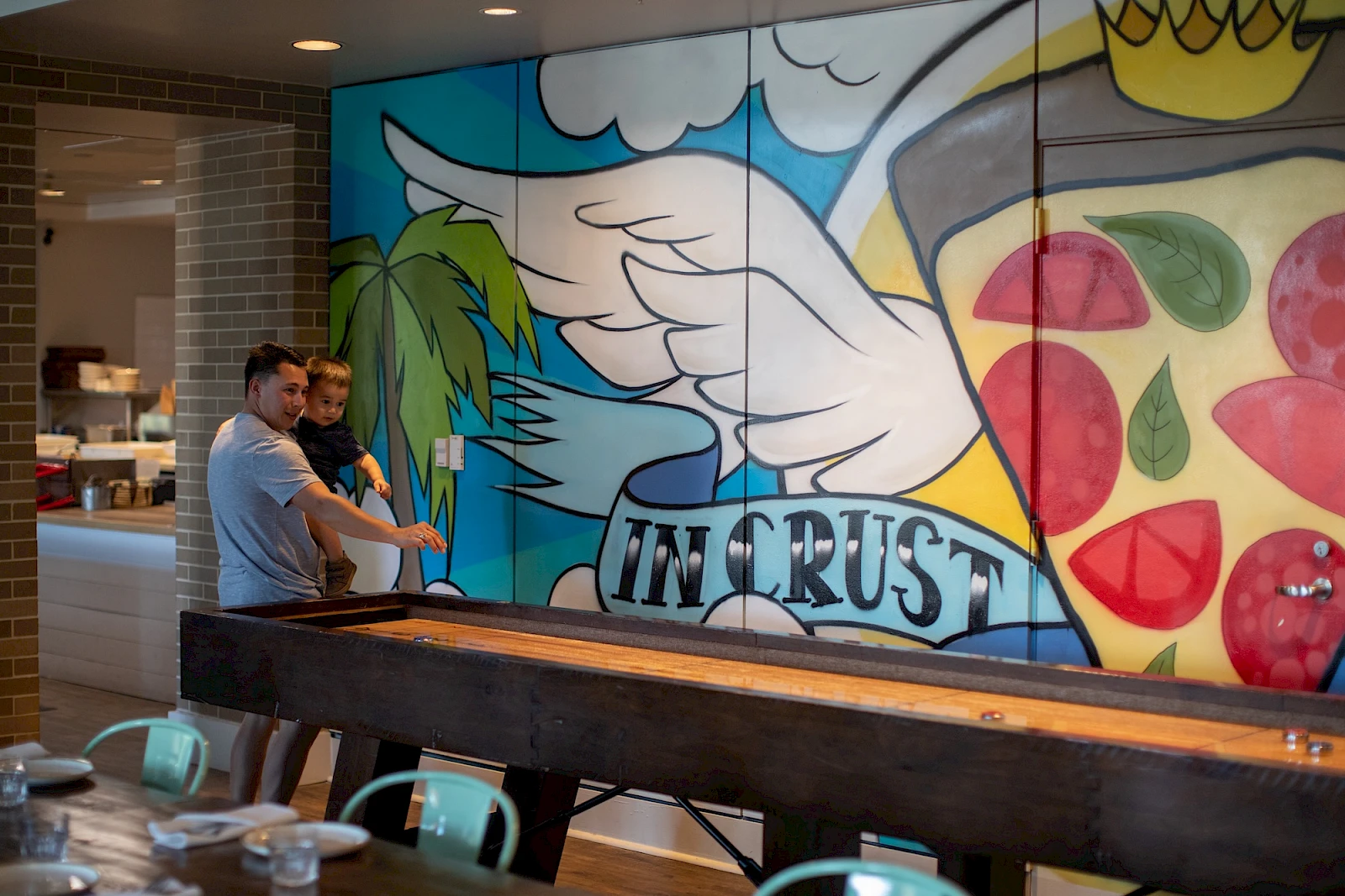 Two people are painting a large colorful mural indoors, featuring wings, a palm tree, a sunlit sky, and the word “IN CRUST” across the center.