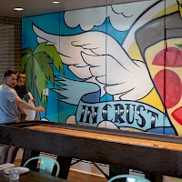 Two people are painting a large colorful mural indoors, featuring wings, a palm tree, a sunlit sky, and the word “IN CRUST” across the center.