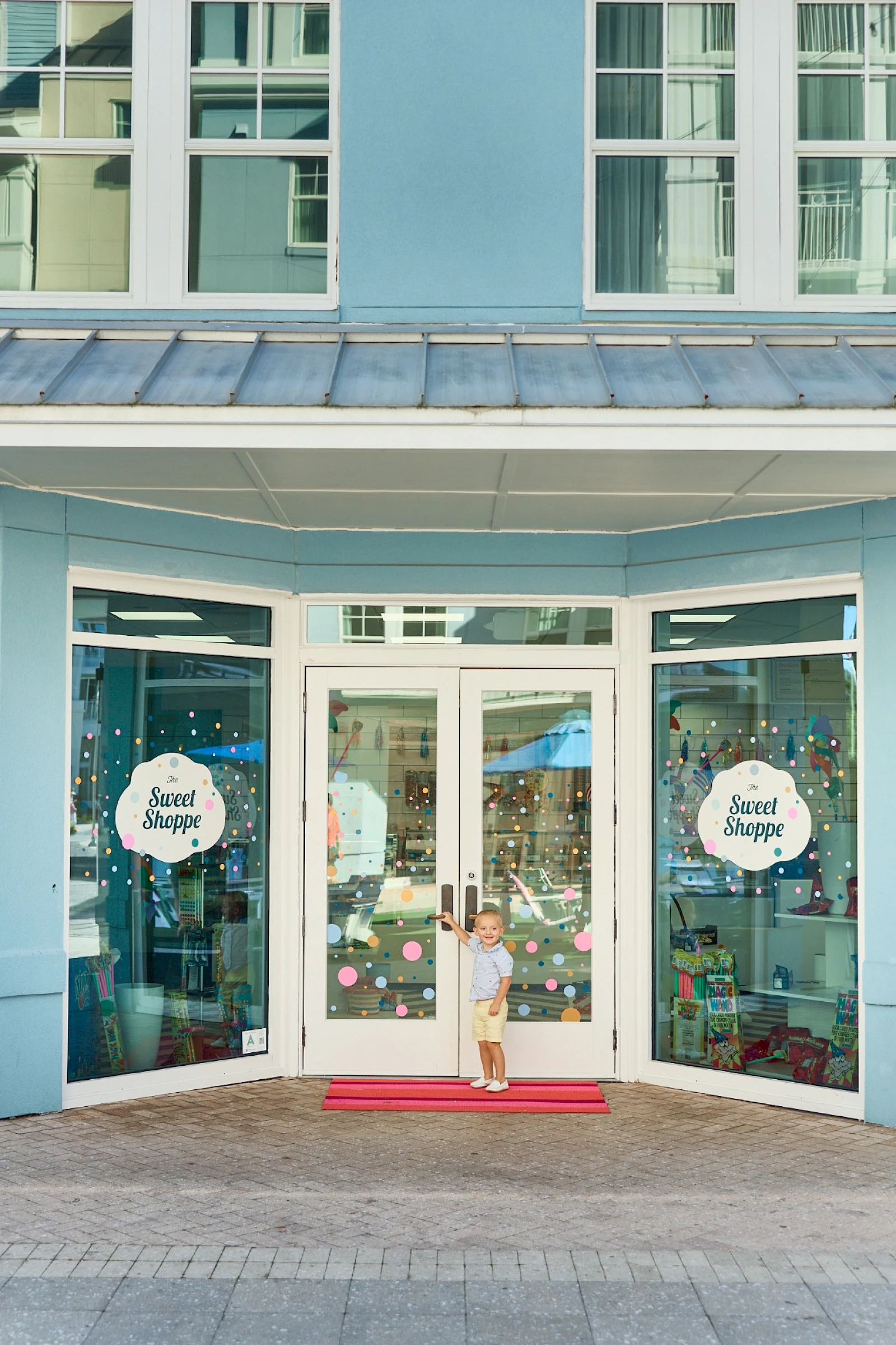 A light blue storefront with open doors, a child standing on a red mat, and circular window signs reading “Sweet Shoppe.”