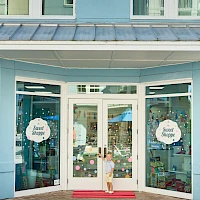 A light blue storefront with open doors, a child standing on a red mat, and circular window signs reading “Sweet Shoppe.”