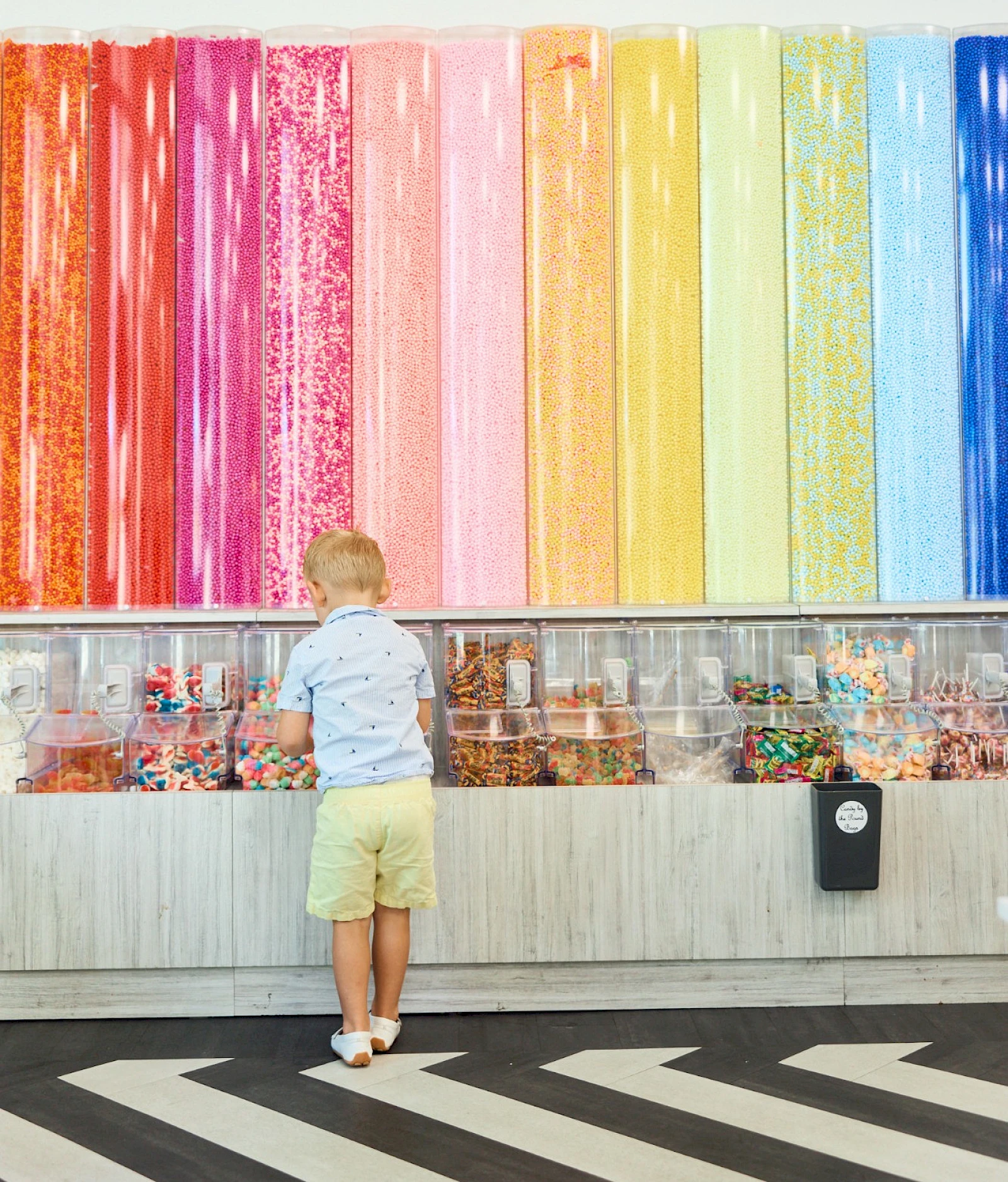 A young child in a light blue shirt and yellow shorts stands facing a colorful wall of candy and art jars, beneath a rainbow mural. End.