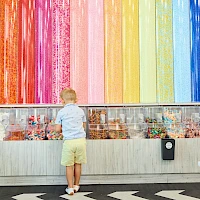 A young child in a light blue shirt and yellow shorts stands facing a colorful wall of candy and art jars, beneath a rainbow mural. End.