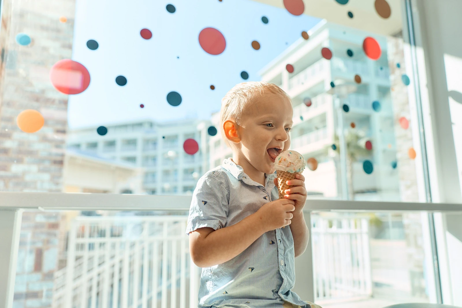 A cheerful toddler enjoys a scoop of ice cream inside a bright, modern cafe, with colorful confetti-like dots decorating the window.
