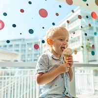 A cheerful toddler enjoys a scoop of ice cream inside a bright, modern cafe, with colorful confetti-like dots decorating the window.