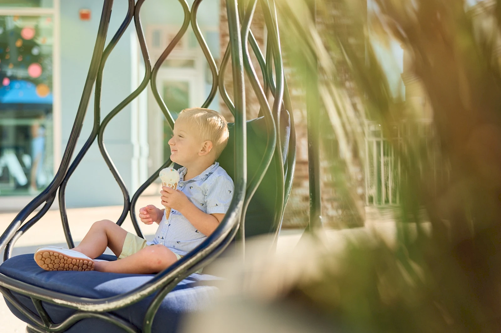 A child sits in a hanging chair outdoors, drinking from a cone-shaped ice cream cup, wearing light blue clothes, with blurred plants and a sunny backdrop.