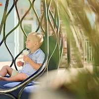 A child sits in a hanging chair outdoors, drinking from a cone-shaped ice cream cup, wearing light blue clothes, with blurred plants and a sunny backdrop.