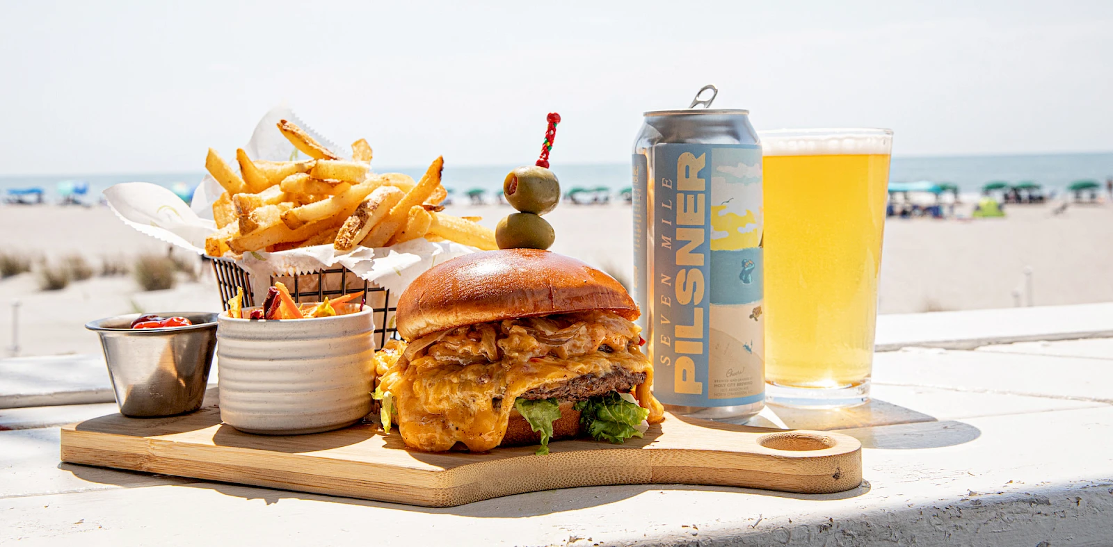 A juicy burger with lettuce and cheese, crispy fries, dipping sauces, a pickle skewer, a beer can, and a glass of beer on a wooden board at the beach.