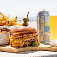 A juicy burger with lettuce and cheese, crispy fries, dipping sauces, a pickle skewer, a beer can, and a glass of beer on a wooden board at the beach.