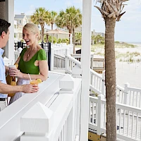Two people chat and toast drinks at an outdoor beachside bar, with palm trees and a sandy shore in the background.