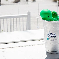 A metal cup labeled “Wild Dunes” with a green toy or paddle inside, on a sunlit outdoor surface near a white fence.