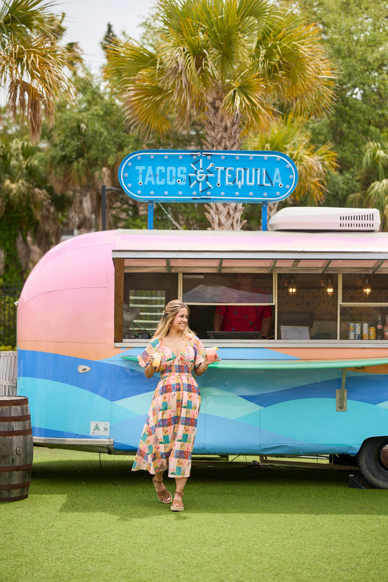 A woman in a patterned dress stands in front of a retro pink-and-blue food truck with a "Tacos Tequila" sign, palm trees in the background.