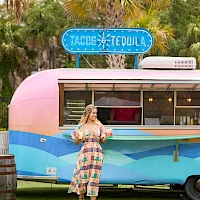 A woman in a patterned dress stands in front of a retro pink-and-blue food truck with a "Tacos Tequila" sign, palm trees in the background.