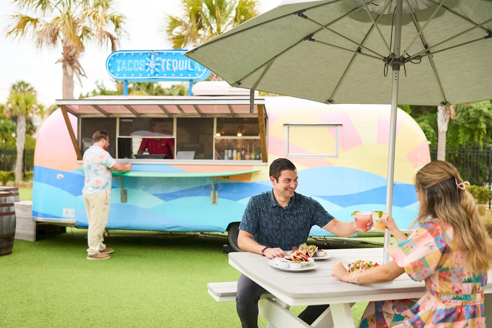 A retro pastel food truck serving tacos and smoothies, outdoors under a big umbrella; two people share a meal at a white picnic table.