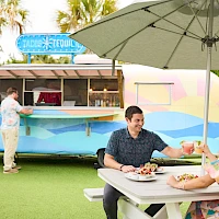 A retro pastel food truck serving tacos and smoothies, outdoors under a big umbrella; two people share a meal at a white picnic table.