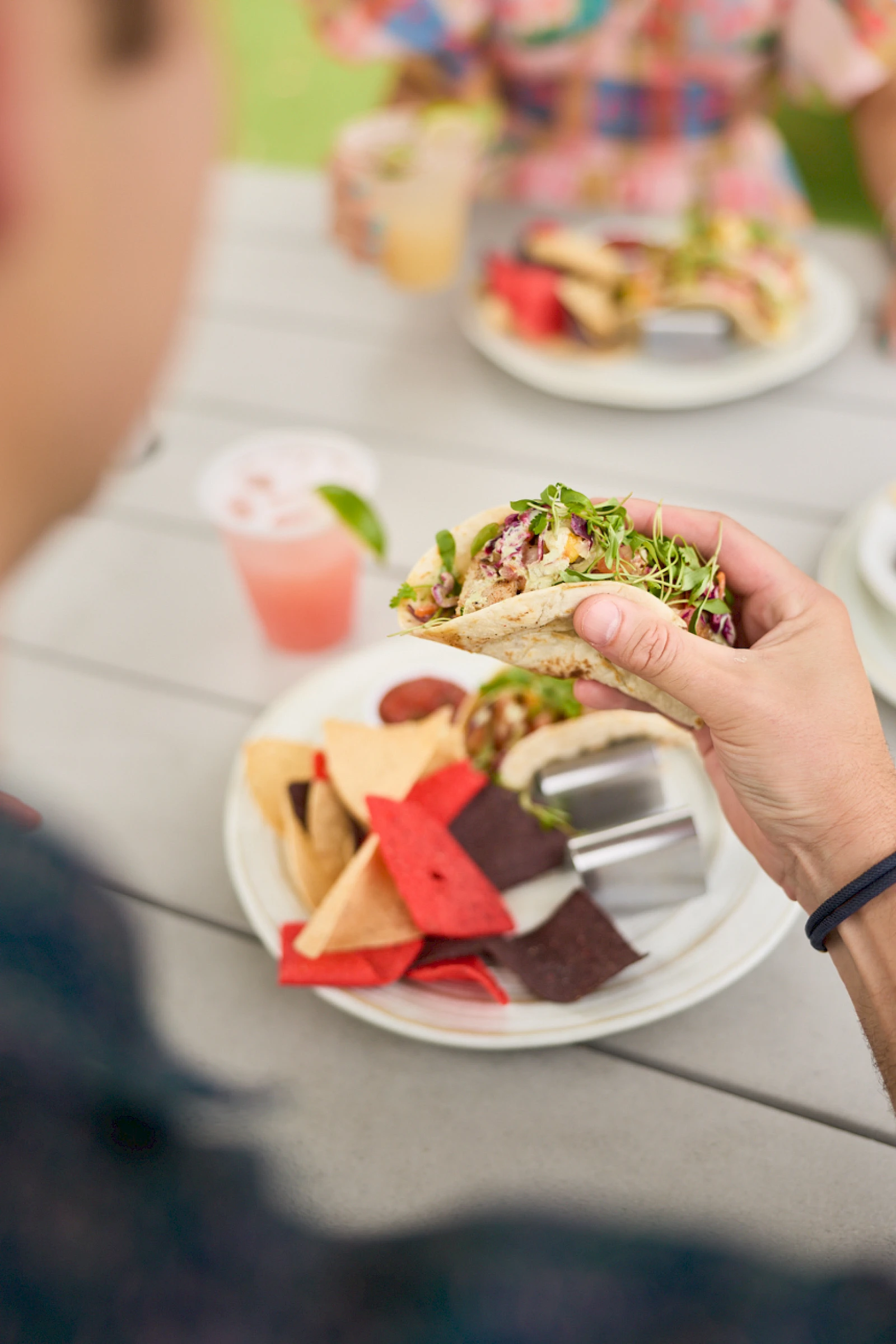 A person holds a veggie-topped sandwich with a plate of colorful chips and a pink drink on a picnic table in the background.