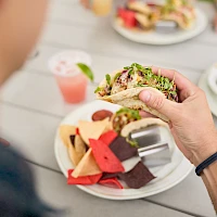 A person holds a veggie-topped sandwich with a plate of colorful chips and a pink drink on a picnic table in the background.