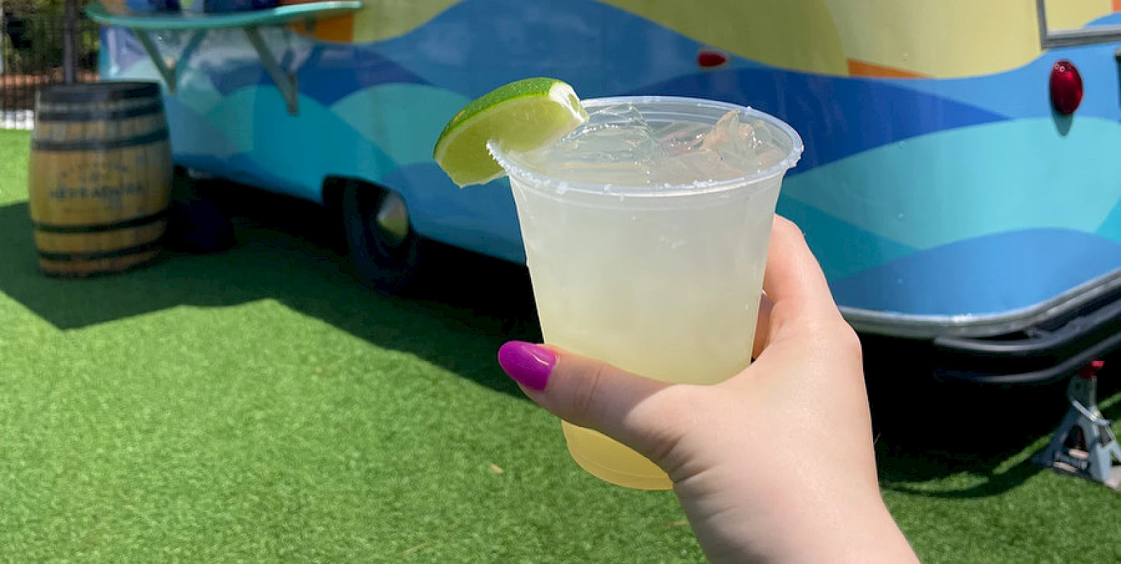 Someone holding a plastic cup with a lime wedge, in front of a colorful bus or trailer parked on grass.