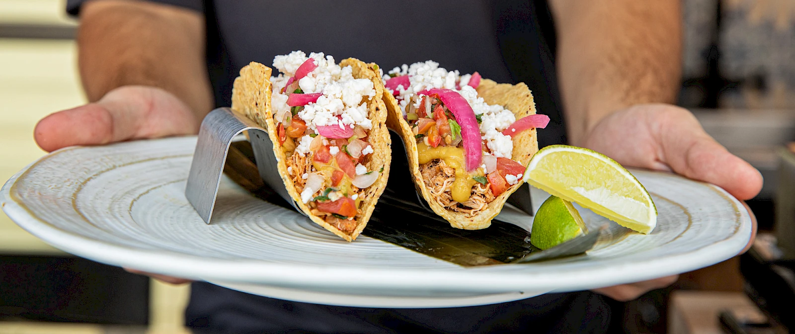 Two tacos filled with vibrant ingredients, topped with crumbled cheese and onions, served on a plate with lime wedges on the side.