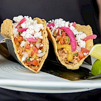 Two tacos filled with vibrant ingredients, topped with crumbled cheese and onions, served on a plate with lime wedges on the side.