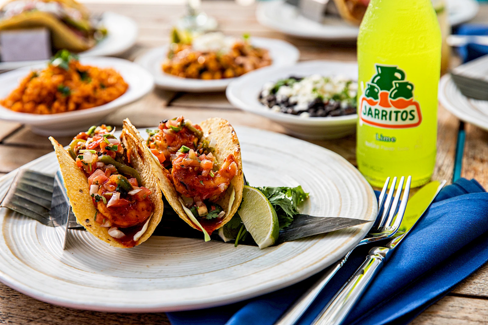 Tacos with beans and salsa on a plate, lime wedge, and a Jarritos lime soda nearby; colorful sides in the background.