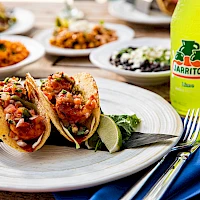 Tacos with beans and salsa on a plate, lime wedge, and a Jarritos lime soda nearby; colorful sides in the background.