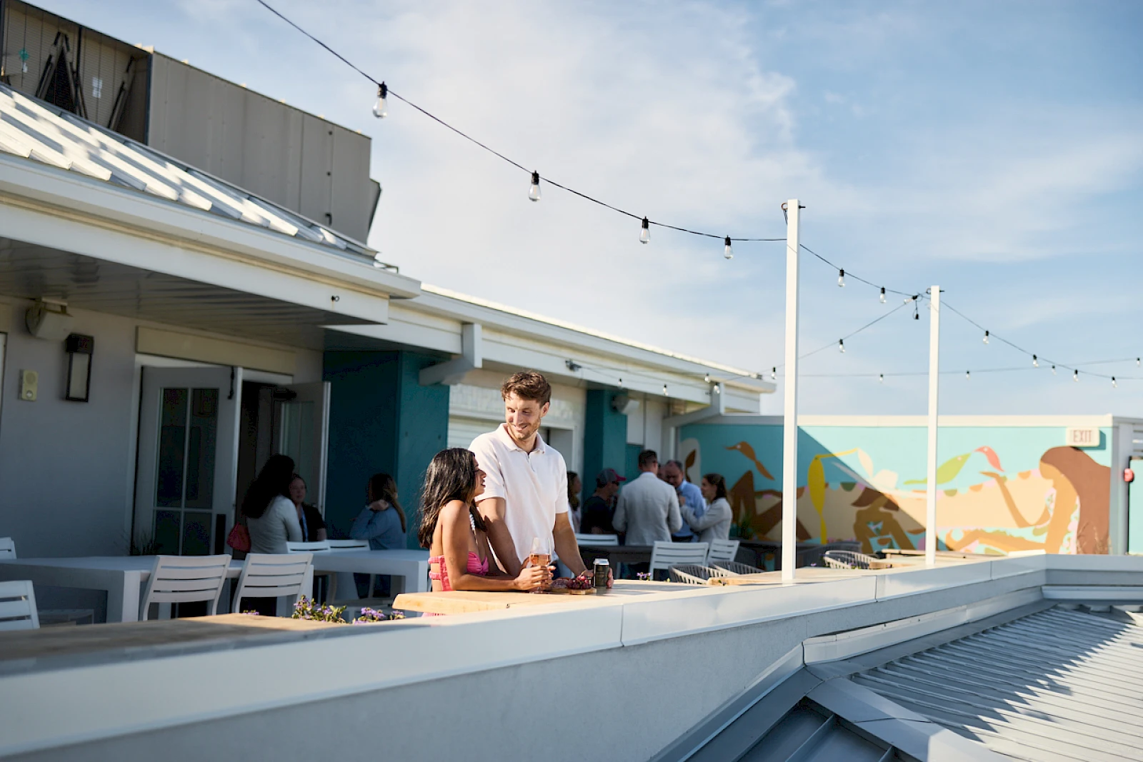 A sunny rooftop hangout with people chatting near a bar, string lights, and a colorful mural in the background.