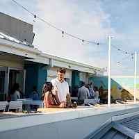 A sunny rooftop hangout with people chatting near a bar, string lights, and a colorful mural in the background.
