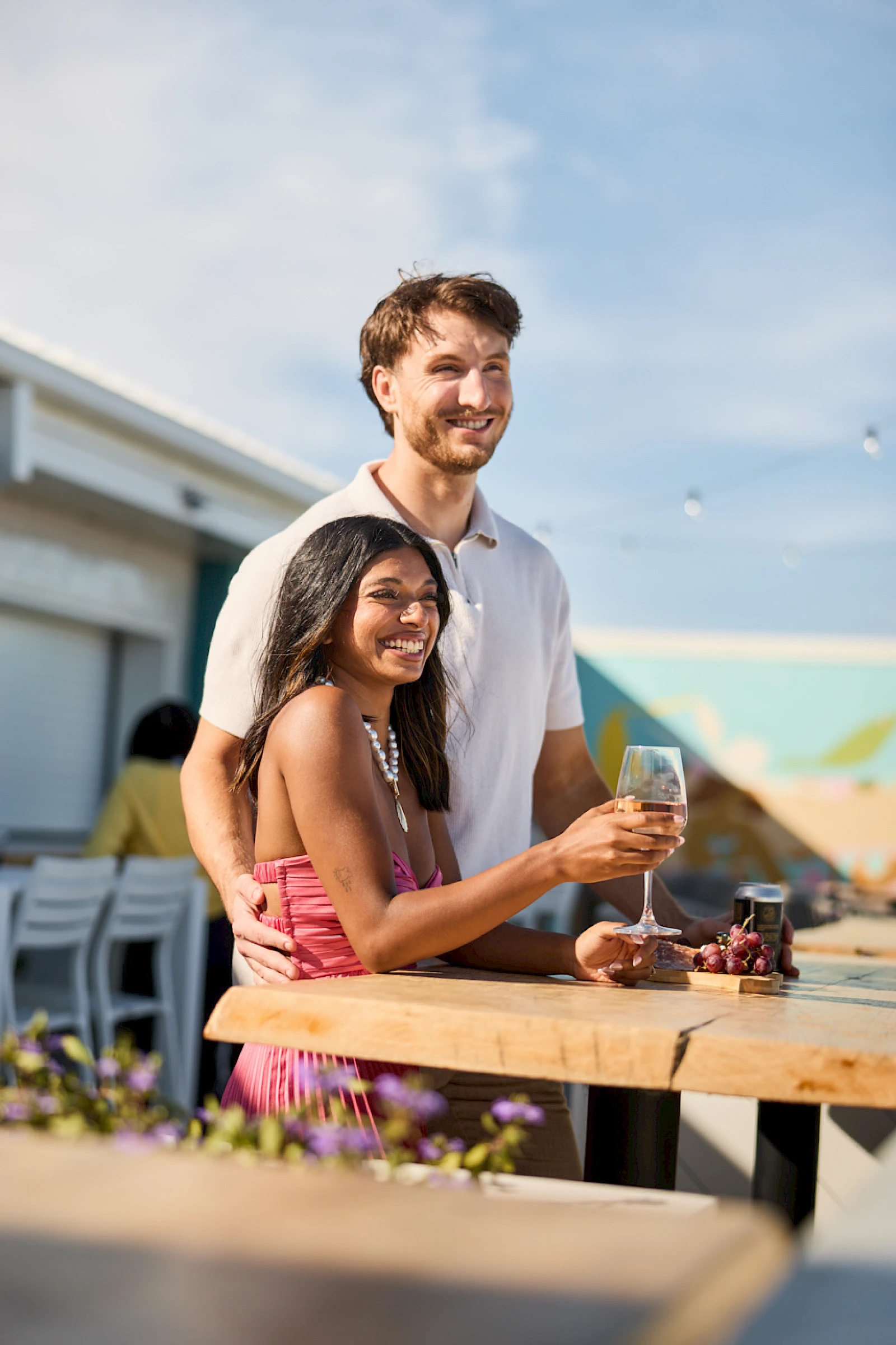 A smiling couple stands by a wooden table with drinks, embracing affectionately at an outdoor cafe or rooftop bar on a sunny day.