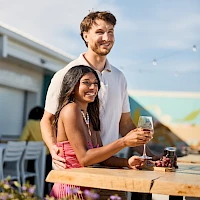 A smiling couple stands by a wooden table with drinks, embracing affectionately at an outdoor cafe or rooftop bar on a sunny day.