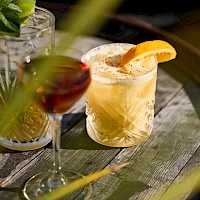 A close-up of three cocktails on a wooden table, garnished with citrus slices and herbs, one with a yellow straw, in a cozy setting.