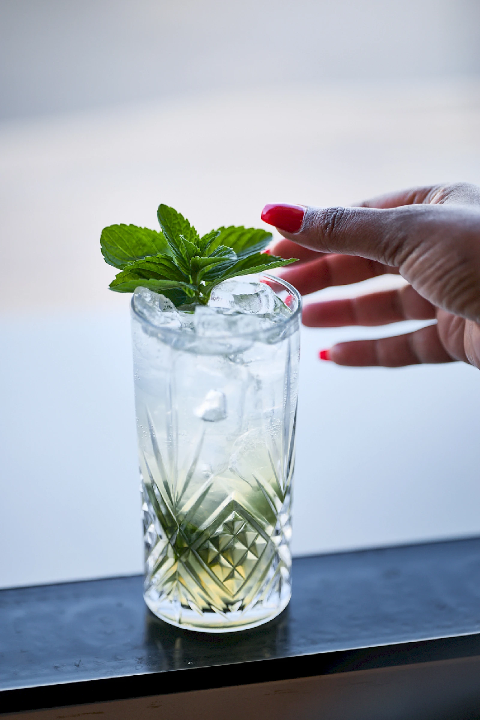 A hand with red nails adds fresh mint garnish to a tall, patterned glass of light-colored drink on a windowsill.