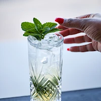 A hand with red nails adds fresh mint garnish to a tall, patterned glass of light-colored drink on a windowsill.
