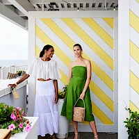 Two women pose on a sunny balcony: one in a white off-shoulder top and lilac skirt, the other in a bright green strapless dress with a tan tote, colorful flowers nearby.