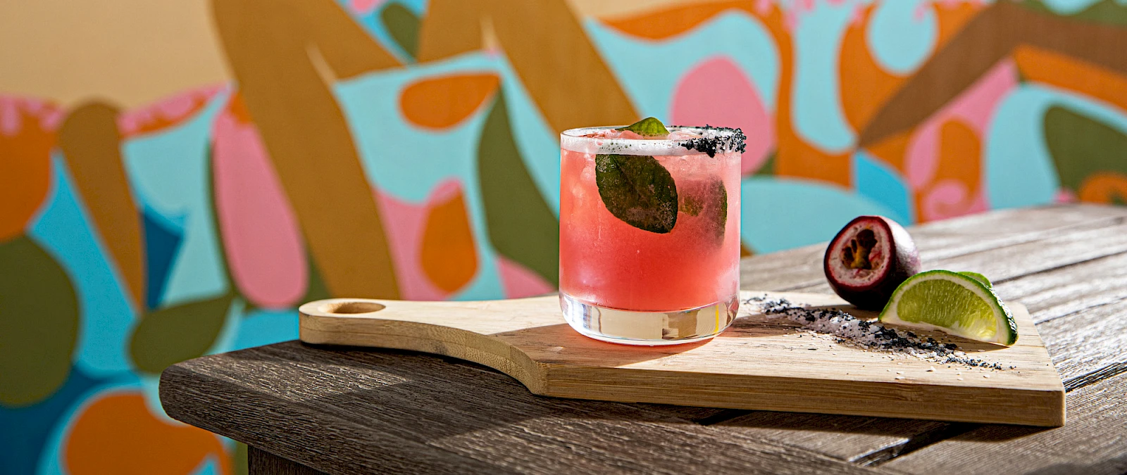 A pink cocktail with a black olive sits on a wooden board, lime wedges and crumbs beside it, against a colorful mural backdrop.